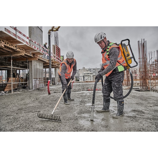 Two construction workers in safety gear are working on a building site. One worker is using a DEWALT DCPS310N concrete vibrator backpack, and the other is leveling wet concrete with a rake. Steel reinforcements and scaffolding are visible in the background.