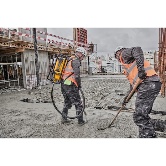 Two construction workers wearing orange safety vests and helmets are working on a concrete floor at a building site. One worker uses a DEWALT backpack-powered tool with a hose, while the other smooths the concrete with a rake.