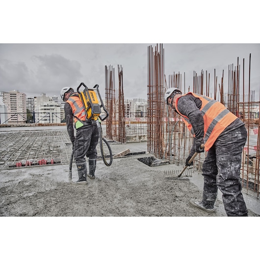 Two construction workers wearing safety helmets and orange reflective vests are working on a concrete surface at a building site. One worker uses a DEWALT backpack tool, while the other uses a rake to spread the concrete. Steel reinforcement bars are visible in the background.