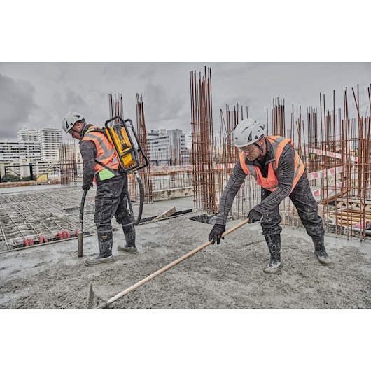 Two construction workers wearing safety helmets and orange vests are working on a concrete slab. One worker uses a DEWALT backpack tool to handle concrete while the other levels the wet concrete with a rake. Reinforcement bars and buildings are visible in the background.