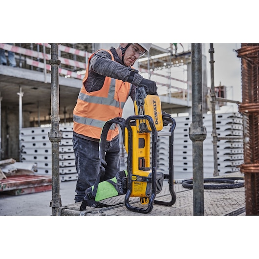 A person wearing a fluorescent safety vest operates a DEWALT DCPS310N power tool on a construction site with scaffolding and concrete elements in the background.