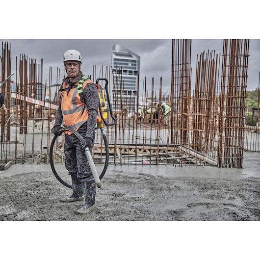 Person wearing a safety helmet and high-visibility vest, operating a DEWALT concrete vibrator tool (DCPS310N) at a construction site with rebar structures and wet concrete.