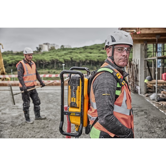 A construction worker wearing a safety vest and helmet is carrying a DEWALT DCPS310N power tool on their back at an outdoor construction site, with another worker seen in the background holding a rake. Faces are blurred for privacy.