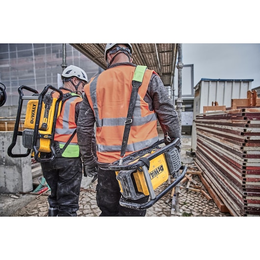 Two construction workers wearing safety helmets and orange reflective vests are walking on a construction site, each carrying a portable DEWALT power tool.