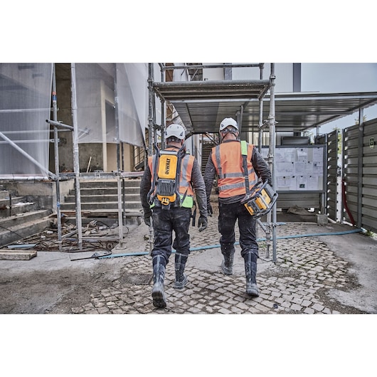 Two construction workers wearing safety helmets and orange reflective vests are walking at a construction site. One worker carries a DEWALT power tool and the other wears a DEWALT backpack. Scaffolding and building materials are visible in the background.