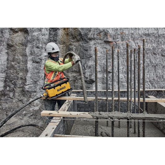 A construction worker wearing safety gear operates a DEWALT portable machine while working with concrete and metal rebar at a building site. The worker is standing on wooden planks and surrounded by foundation forms and exposed rebar.