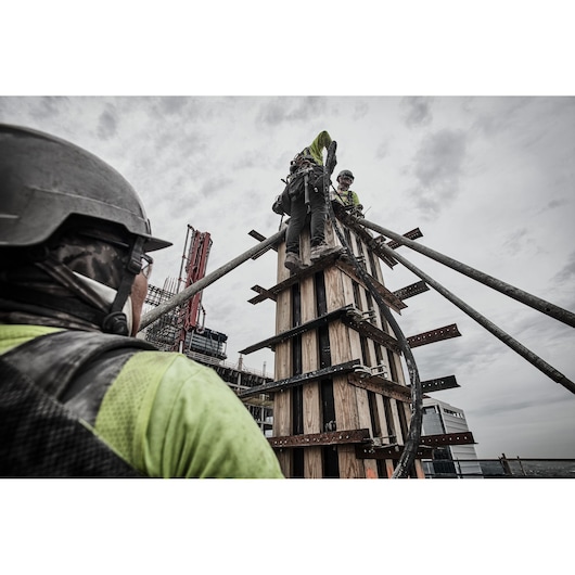Several construction workers, wearing safety gear including helmets and high-visibility clothing, operate on a tall wooden structure at a construction site. The workers are using equipment and hoses, and the scene shows industrial activity with overcast skies.