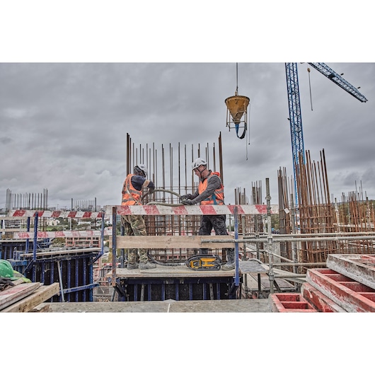 Two construction workers wearing helmets and safety vests are working on a building site with metal rebar structures. A DEWALT tool is placed on the platform in front of them. There is construction equipment, scaffolding, and a blue crane in the background under a cloudy sky.