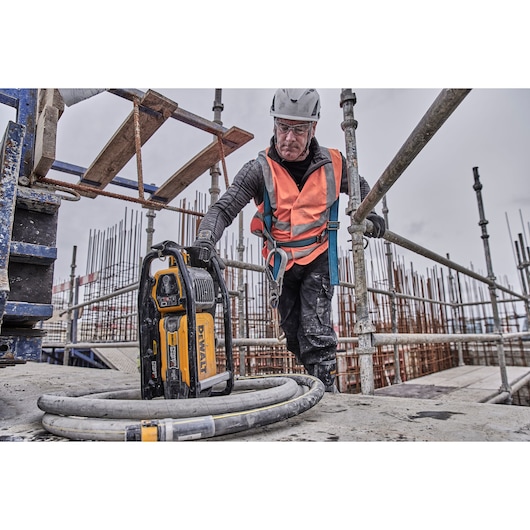 A DEWALT DCPS320N power tool is placed on a construction site platform, with a person wearing safety equipment and an orange vest standing nearby. The environment includes scaffolding, exposed rebar, and construction materials.