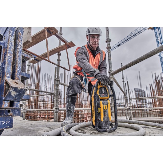 A construction worker wearing safety gear and an orange vest uses a DEWALT DCPS320N tool at an active construction site with scaffolding and building materials visible in the background.