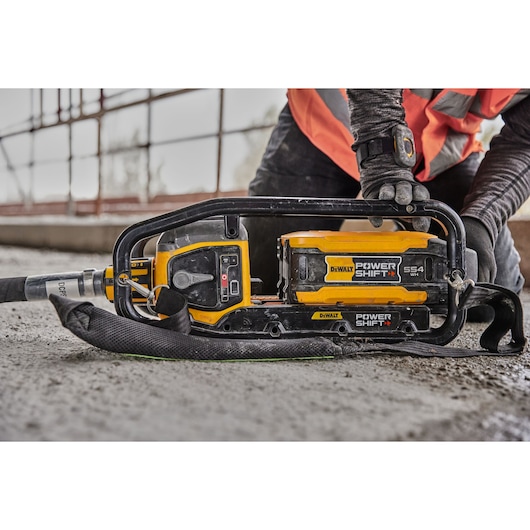 A close-up of a DEWALT DCPS320N Power Shift concrete vibrator tool resting on a concrete surface at a construction site. A person wearing an orange safety vest and gloves is adjusting or handling the tool.