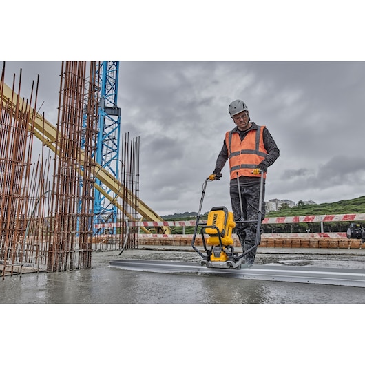 A construction worker wearing a safety helmet and orange reflective vest operates a DEWALT machine to smooth concrete at a construction site, surrounded by metal rebars and construction equipment.