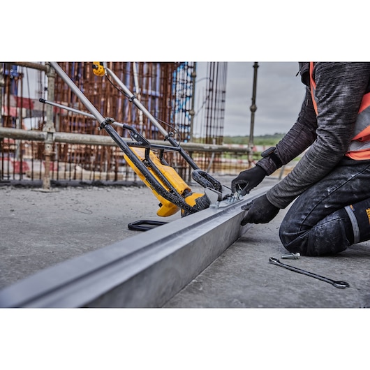 A person wearing work gloves and safety gear is assembling or adjusting a DEWALT power tool on a construction site. The tool is positioned alongside a metal beam, with other construction equipment and scaffolding visible in the background.