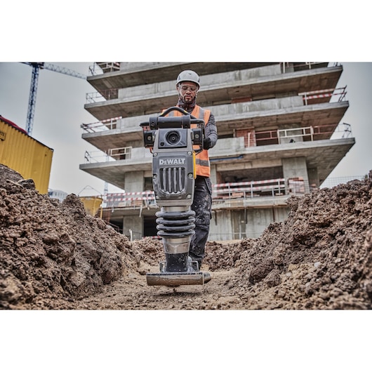 A construction worker wearing a helmet and high-visibility vest operates a DEWALT compactor on a muddy site with a partially built multi-story structure in the background.