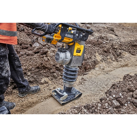 A DEWALT DCPS660N rammer compactor being operated on a construction site, compacting soil between areas of earth and gravel.