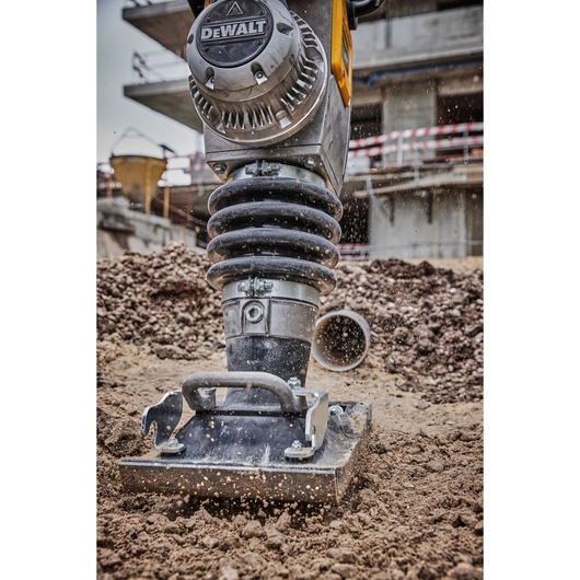 A close-up view of a DEWALT compactor tool in action on a construction site, compacting soil with dust particles flying around.