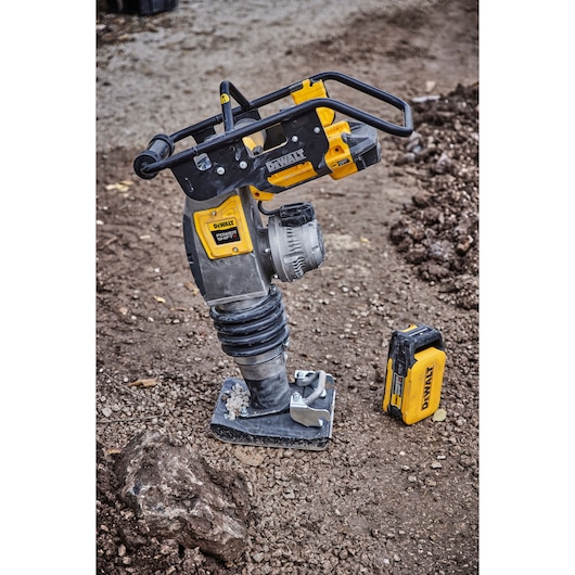 A DEWALT cordless rammer for soil compaction is shown standing on a gravel surface, with a yellow and black battery placed beside it. The machine features a sturdy handle and spring mechanism, and is designed for construction or landscaping work.