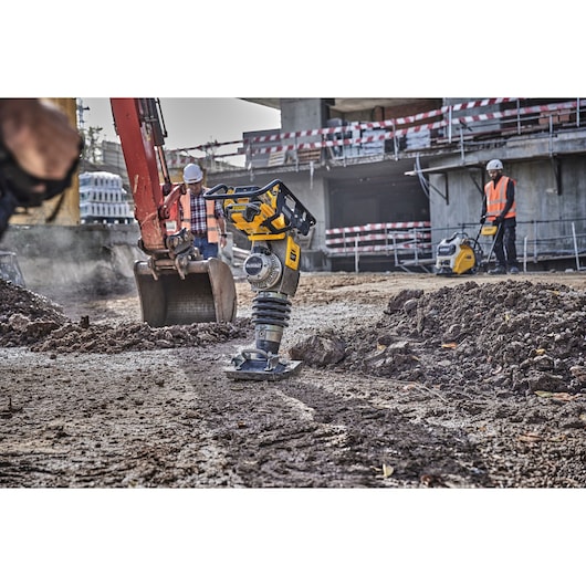 A DEWALT DCPS660N power tool is being used on a construction site to compact soil. Workers in safety gear and construction equipment are visible in the background.