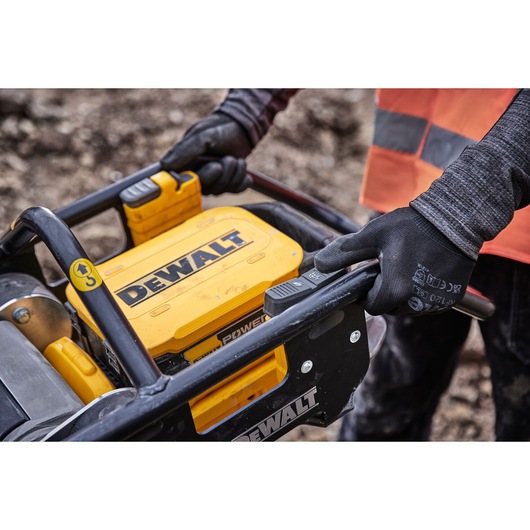 Close-up of a person wearing gloves operating a DEWALT power tool outdoors. The DEWALT branding is clearly visible on the yellow machine.