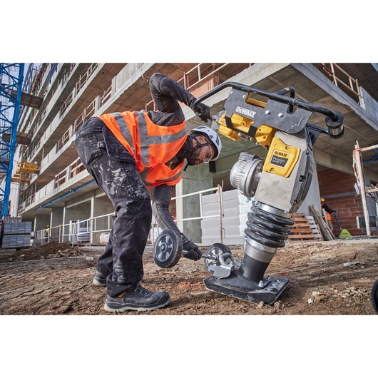 A construction worker wearing a safety helmet and orange reflective vest is using a DEWALT power tool on a construction site. The worker is operating the DEWALT tool on bare ground near a large building under construction.
