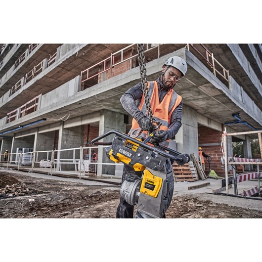 A person wearing safety gear uses a DEWALT DCPS660N power tool at a construction site in front of a partially built concrete building.