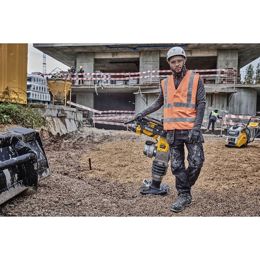 A construction worker wearing a safety helmet and orange reflective vest is operating a DEWALT DCPS660N power tool on a gravel surface at a construction site, with concrete structures and other workers in the background.