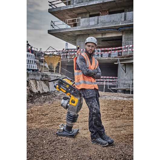 A construction worker wearing a safety helmet and orange high-visibility vest stands with arms crossed next to a DEWALT DCPS660N power tool on a construction site with a partially built building in the background.