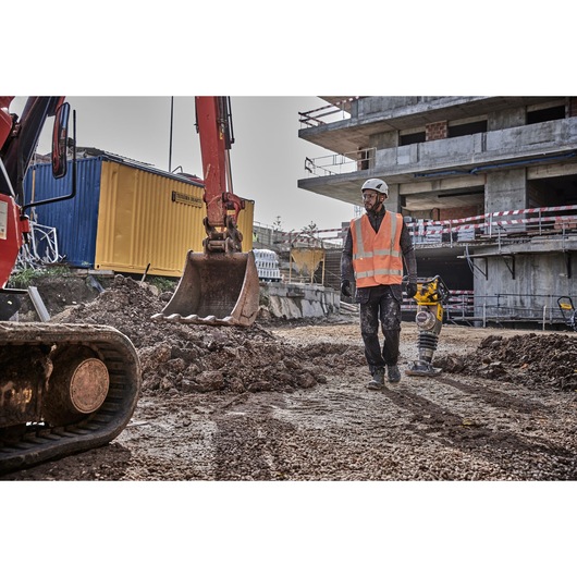 A construction worker wearing a safety helmet and orange high-visibility vest operates a yellow DEWALT power tool on a dirt surface at a construction site. An excavator and a yellow shipping container are visible in the background, along with a partially built concrete structure.