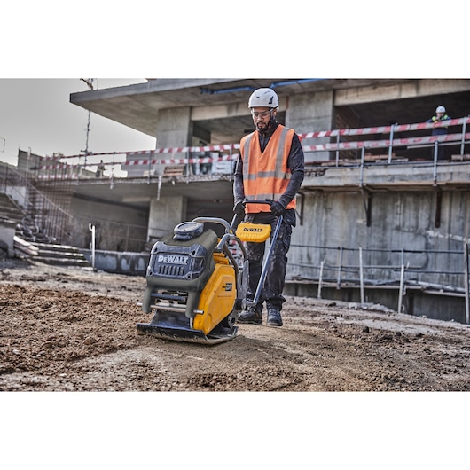 A construction worker wearing a safety helmet and orange reflective vest operates a yellow DEWALT plate compactor on a dirt surface at an outdoor construction site with an unfinished concrete building in the background.