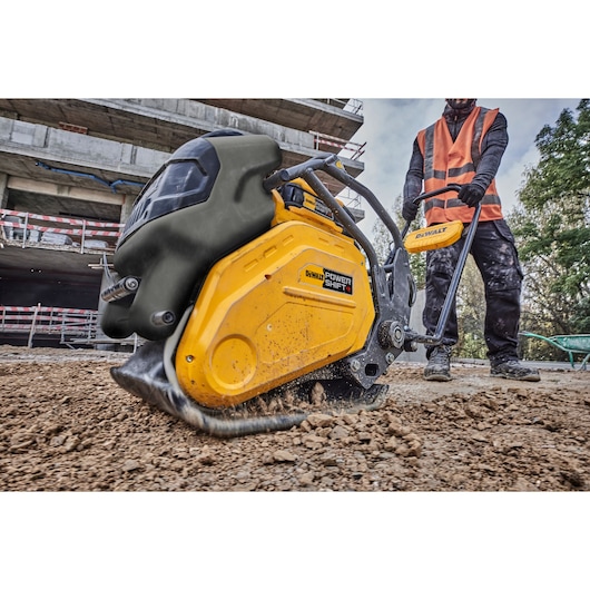 A DEWALT Power Shift plate compactor being operated on a construction site, compacting soil. A person wearing a hi-vis vest and work boots is guiding the machine.