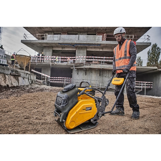 A construction worker wearing safety gear and a reflective vest operates a yellow DEWALT plate compactor (model DCPS7154N) on a construction site with a partially built concrete structure in the background.