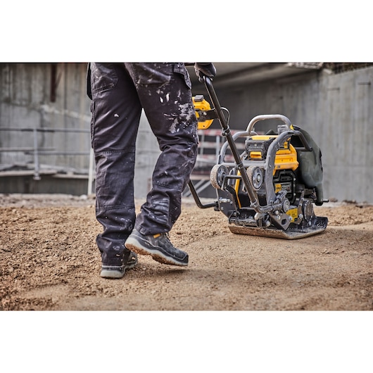A person operating a DEWALT plate compactor on a construction site, pressing soil or gravel with the machine.