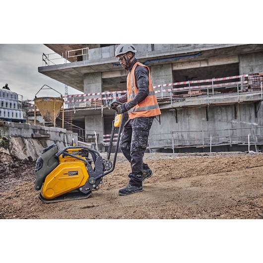 A construction worker wearing safety gear operates a DEWALT plate compactor on a building site, with an unfinished concrete structure in the background.