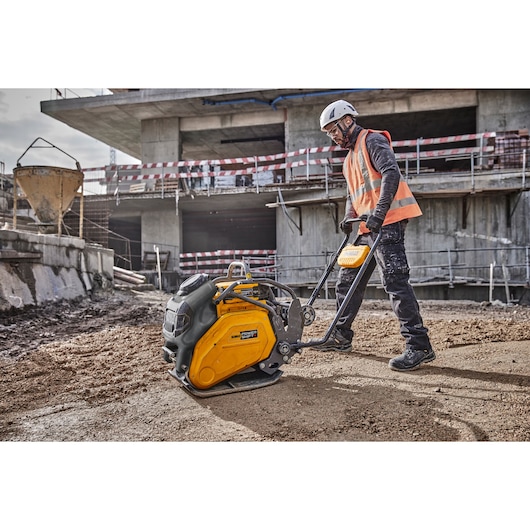 A worker wearing a safety helmet and orange vest operates a yellow and black DEWALT compactor machine on a construction site, with a partially constructed building in the background.