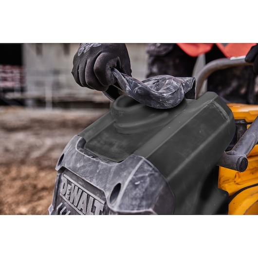A close-up view of a person wearing gloves and opening the cover of a DEWALT power tool, with construction materials and equipment in the background.