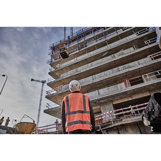 A construction worker wearing a high-visibility orange vest and a white helmet stands in front of an unfinished concrete building at a construction site. A piece of equipment is being lifted by a crane. Other workers and construction materials are visible in the background.