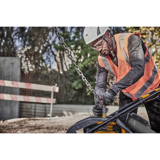 A construction worker wearing a high-visibility orange vest and white safety helmet is operating a DEWALT tool outdoors. The worker is connecting a heavy-duty chain to the equipment, with protective gloves and safety barriers in the background.