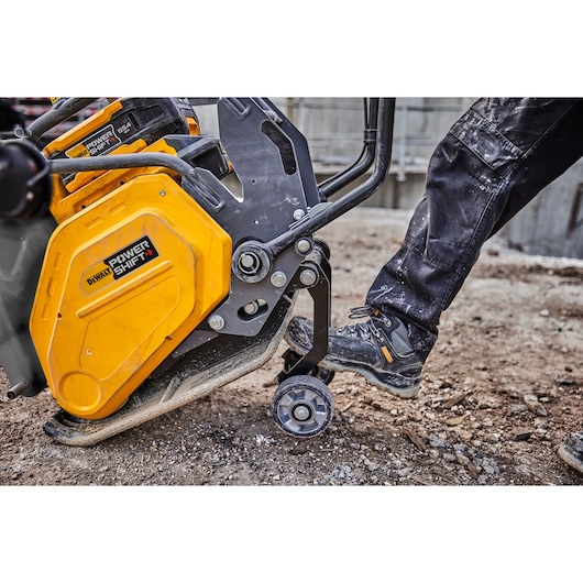 Close-up of a construction worker operating a yellow DEWALT Power Shift machine on a rough outdoor surface. The worker's foot is positioned on a wheel lever attached to the equipment.