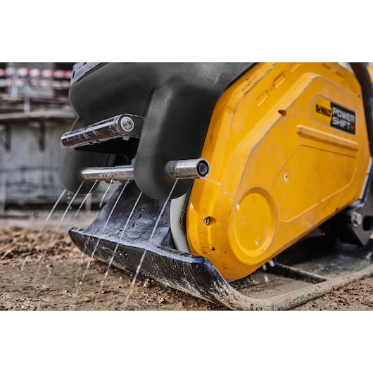 Close-up of a DEWALT Power Shift plate compactor in use, with water spraying from metal tubes onto the ground.