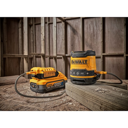 A DEWALT DCR009 portable jobsite Bluetooth speaker connected to a DEWALT POWERSTACK 18V battery, both placed on a wooden surface in a construction site setting.