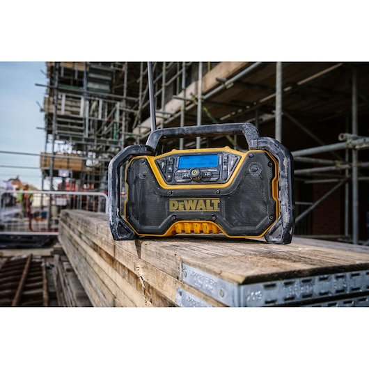 A DEWALT portable radio, model DCR029, is placed on a stack of wooden planks at a construction site. The radio has a rugged black and yellow design and a handle on top, with scaffolding and construction materials visible in the background.