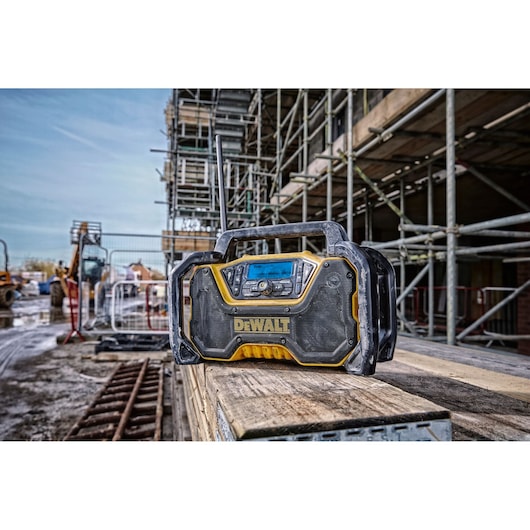 A DEWALT DCR029 rugged portable worksite radio with a digital display, placed on wooden planks at a construction site with scaffolding and equipment in the background.