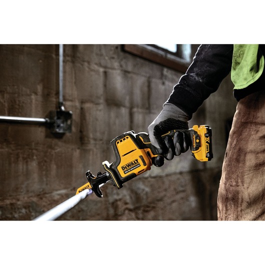 Person uses a DEWALT DCS312NT cordless reciprocating saw, cutting a pipe indoors with gloves.