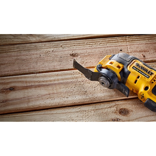 A DEWALT cordless oscillating multi-tool is shown cutting into wooden planks, creating sawdust on the surface.