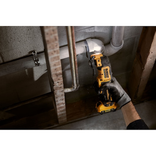 A person wearing a work glove uses a DEWALT cordless oscillating multi-tool to cut a white PVC pipe in a utility space with exposed pipes and wood framing.