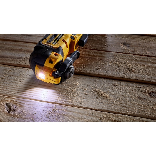 A DEWALT cordless oscillating multi-tool with a built-in LED light is shown in action on a wooden surface with wood dust around.