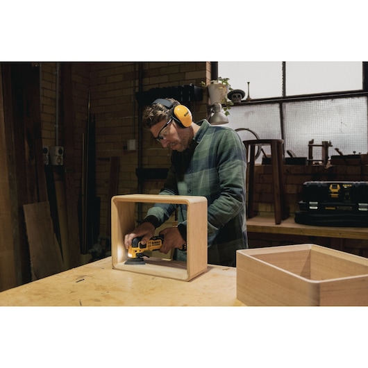 Person sanding wooden box with a DEWALT tool in a workshop wearing safety gear.