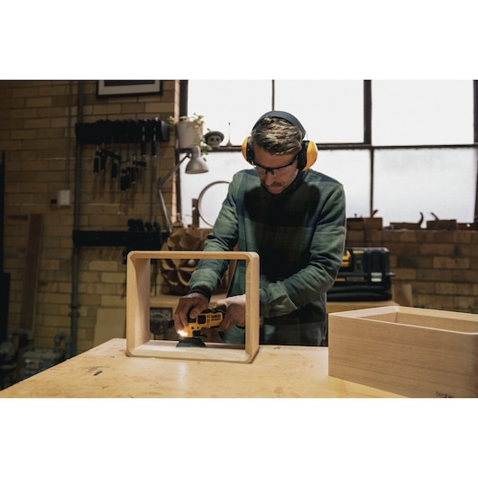 Person using a DEWALT sander on a wooden frame in a workshop, wearing ear and eye protection.