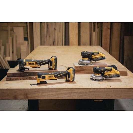 Four DEWALT cordless power tools on a wooden table in a workshop, including sanders and oscillating multi-tools.