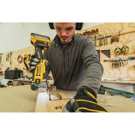 Person uses a DEWALT DCS359N-XJ cordless saw to cut wood on a workbench in a workshop.